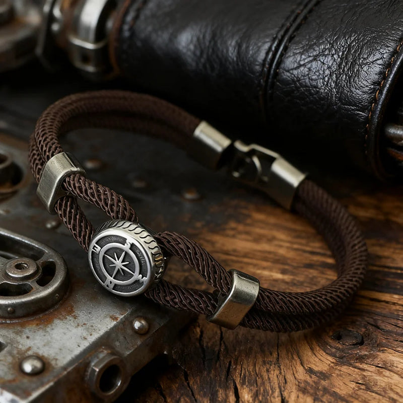 Brown braided bracelet with a silver compass charm on a wooden surface with leather and metal elements.