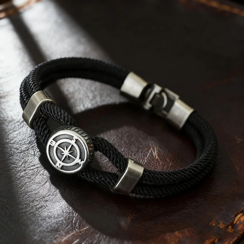 Black braided bracelet with silver compass-like clasp on a dark leather surface