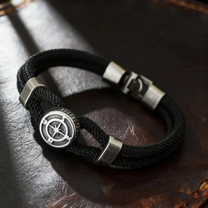 Black braided bracelet with silver compass-like clasp on a dark leather surface