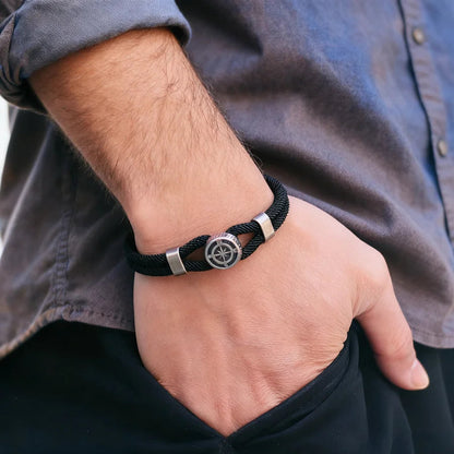 Close-up of a hand wearing a black braided bracelet with a silver compass charm.